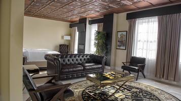 Habitación de hotel con sofá de cuero con capitonado, una mesa redonda, una cama, una silla, un ventanal con cortinas y un techo decorativo.