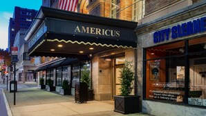 Un teatro con una bandera estadounidense, un cartel luminoso que dice 'AMERICUS' y una tienda llamada 'CITY CLEAN'.