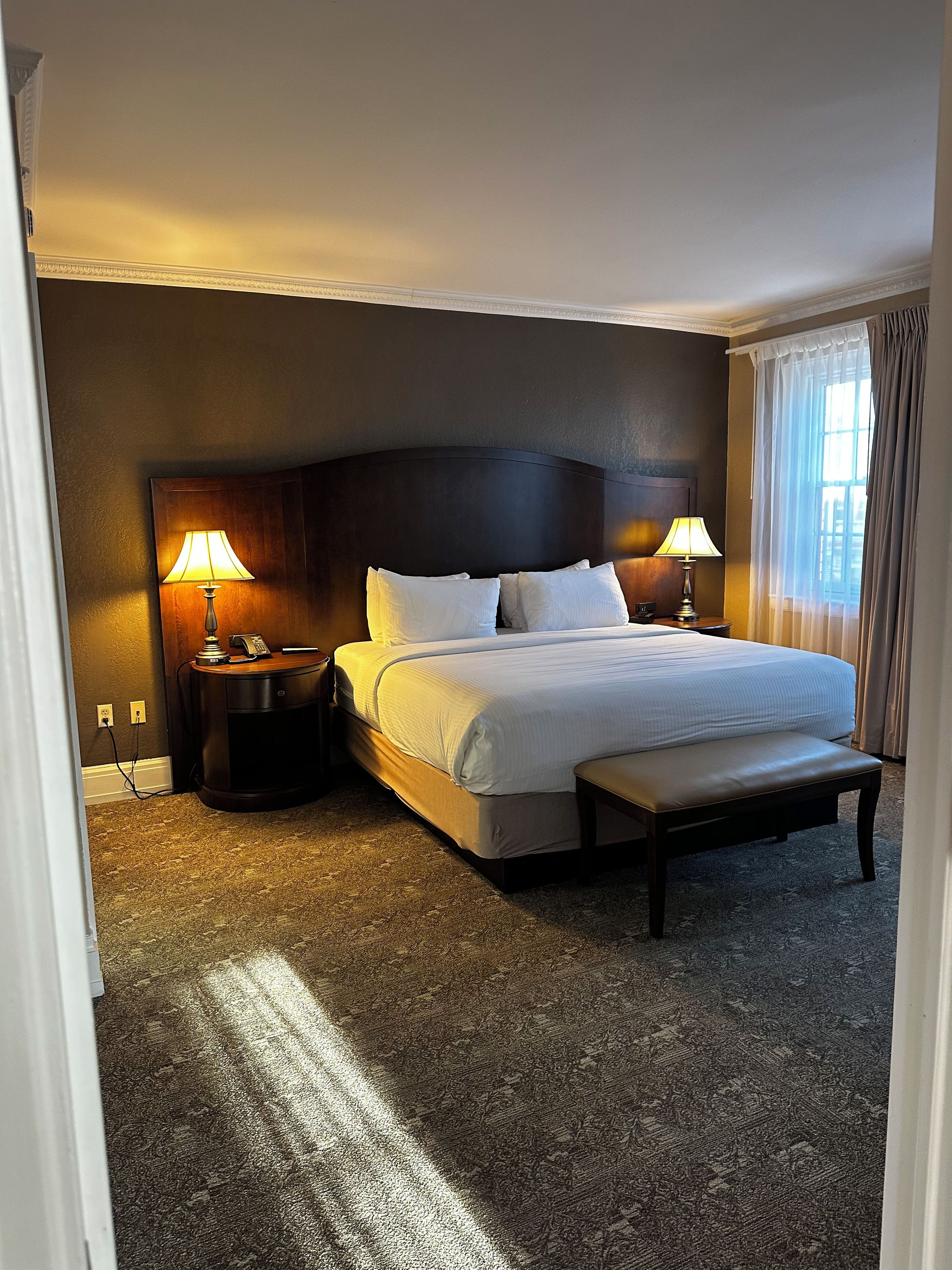 A hotel room with a large bed, two bedside tables with lamps, a bench at the foot of the bed, and a carpeted floor.