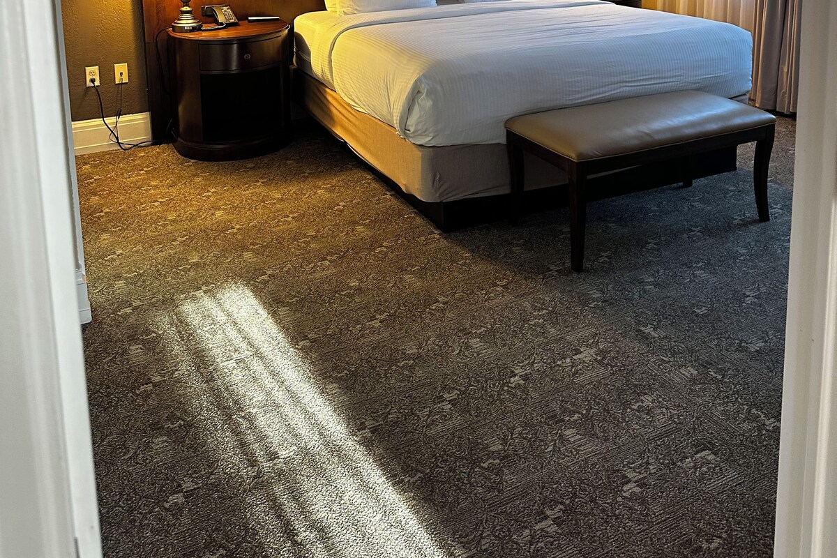 A hotel room with a large bed, two bedside tables with lamps, a bench at the foot of the bed, and a carpeted floor.