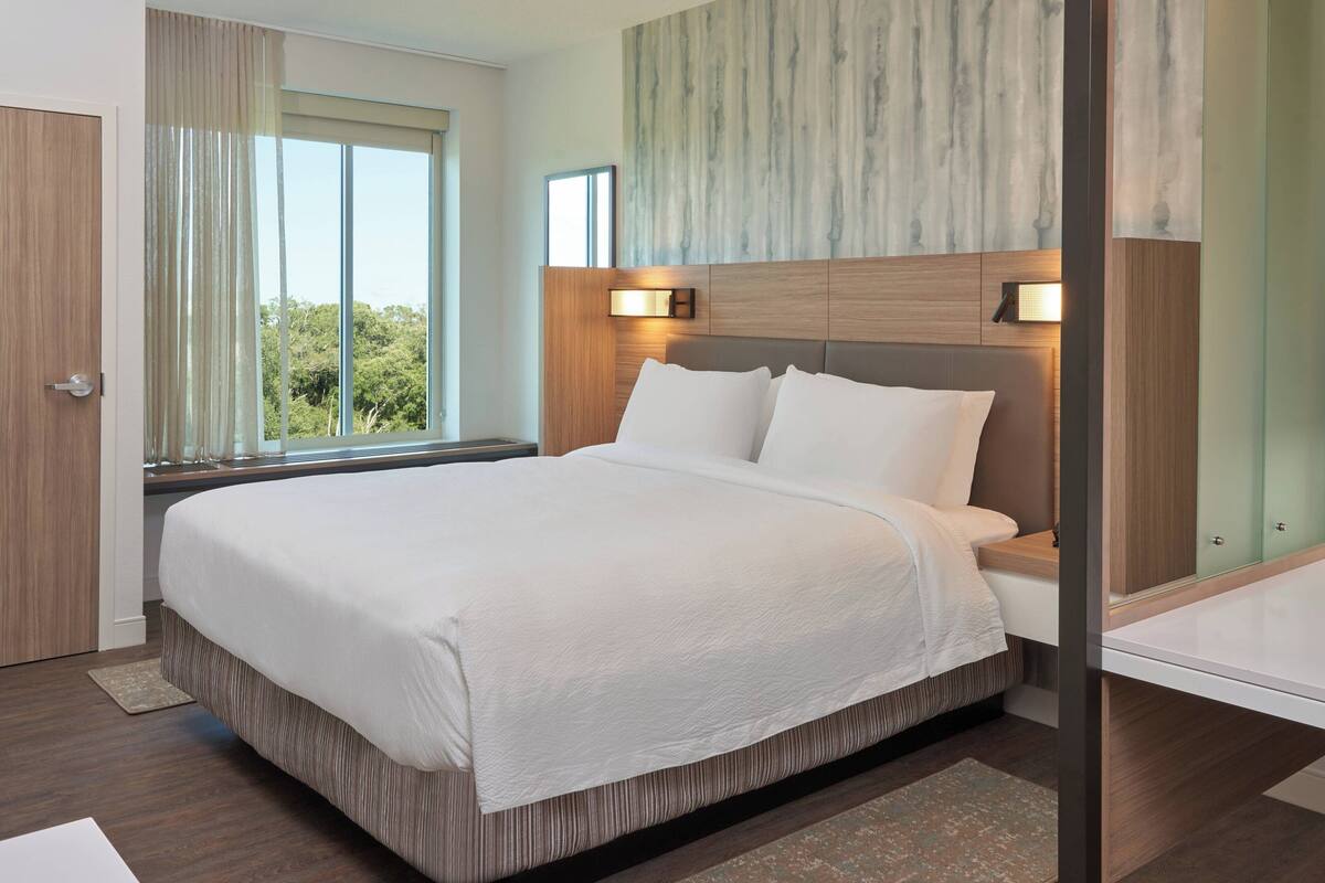 Ein modernes Hotelzimmer mit einem großen Bett, einem hölzernen Kopfteil und Blick auf Bäume durch das Fenster.