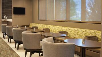 A hotel room with a seating area featuring round tables and chairs, a flat-screen TV mounted on the wall, and a large window with a view of trees.