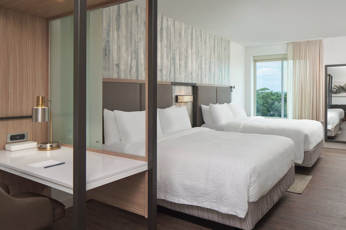 Ein Hotelzimmer mit zwei Betten, einem Schreibtisch, einem Stuhl, einer Lampe und einem großen Fenster mit Vorhängen.