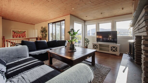 A spacious living room with a wooden ceiling, large windows, a flat-screen TV, and a stone fireplace. - Penthouse Apartment (Bergen)