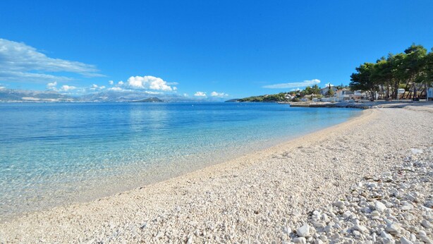 Una spiaggia di ciottoli con acque cristalline, una catena montuosa in lontananza e una fila di case lungo la riva.