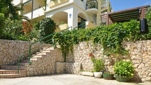 Una scala in pietra conduce a un edificio con facciata bianca e ringhiere verdi. Vasi con piante adornano il muro in pietra e, sullo sfondo, si vede un pergolato in legno.