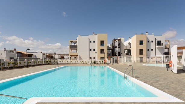 A swimming pool with a tiled deck, surrounded by modern buildings with balconies and a clear sky. - Apartamento con Terraza y piscina Pueblo Majorero (Caleta de Fuste - Antigua)