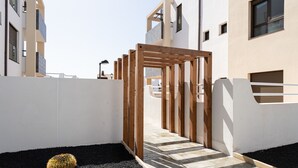 A modern building with a wooden pergola entrance, a black gravel path, and a cactus in the foreground. - Apartamento con Terraza y piscina Pueblo Majorero (Caleta de Fuste - Antigua)