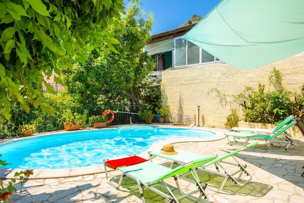 A swimming pool with lounge chairs and a lifebuoy, surrounded by greenery and a building.