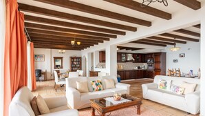 A spacious living area with white sofas, a wooden coffee table, and a kitchen in the background.