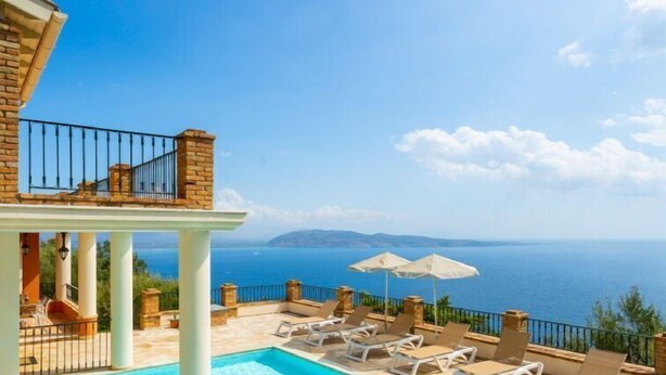 A pool with clear blue water, surrounded by lounge chairs and umbrellas, overlooking a scenic view of the sea and distant mountains.