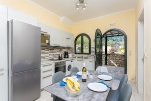 Cozinha compacta com geladeira, fogão e máquina de lavar. Há uma mesa de jantar com cadeiras e uma tigela de bananas.