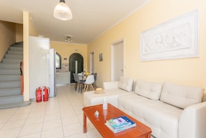 Sala de estar com sofá, mesa de centro e área de jantar com cadeiras e mesa. Há uma escada e uma porta que dá para outro cômodo.