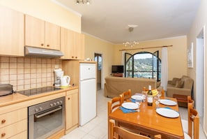 Une cuisine compacte avec des meubles en bois, un four encastré et un réfrigérateur. Une table à manger avec des chaises est placée au centre, offrant une vue sur un balcon et le paysage extérieur.