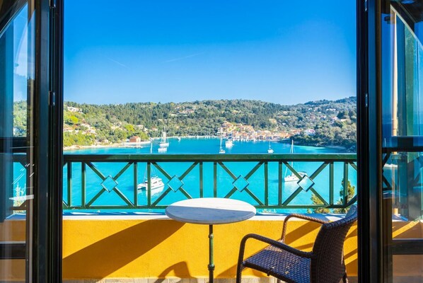A balcony with a table and chair, overlooking a turquoise bay with boats and a coastal town.