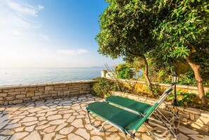 Une terrasse pavée de pierres, avec deux fauteuils de jardin verts et un mur de pierres donnant sur une étendue d’eau.