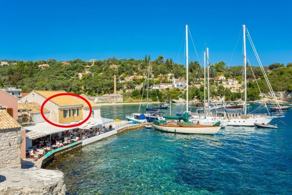 Una marina costera con barcos amarrados, un edificio de piedra con fachada amarilla y una colina con casas al fondo.