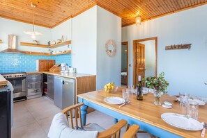 A modern kitchen with a dining area, featuring a wooden ceiling, blue tiled backsplash, stainless steel appliances, and a wooden dining table set for four. - Limani - Two Bedroom Apartment, Sleeps 4 (Longos)