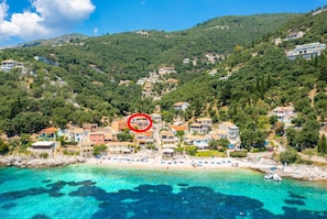 A coastal village with houses on a hillside, a beach with sun loungers, and clear turquoise waters.