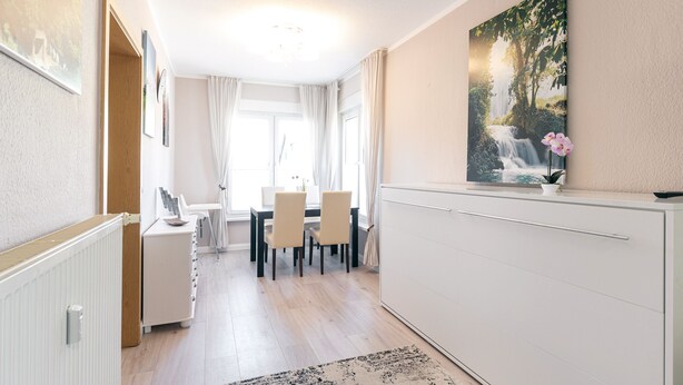 A modern dining area with a white table and chairs, a white cabinet with a painting, and a view of a waterfall through the window. - Vacation apartment Waldesruh (Thale)