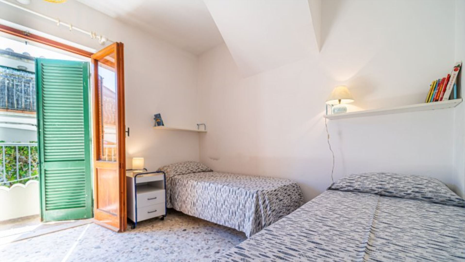 A small, bright room with two beds, a desk, and a window with green shutters.