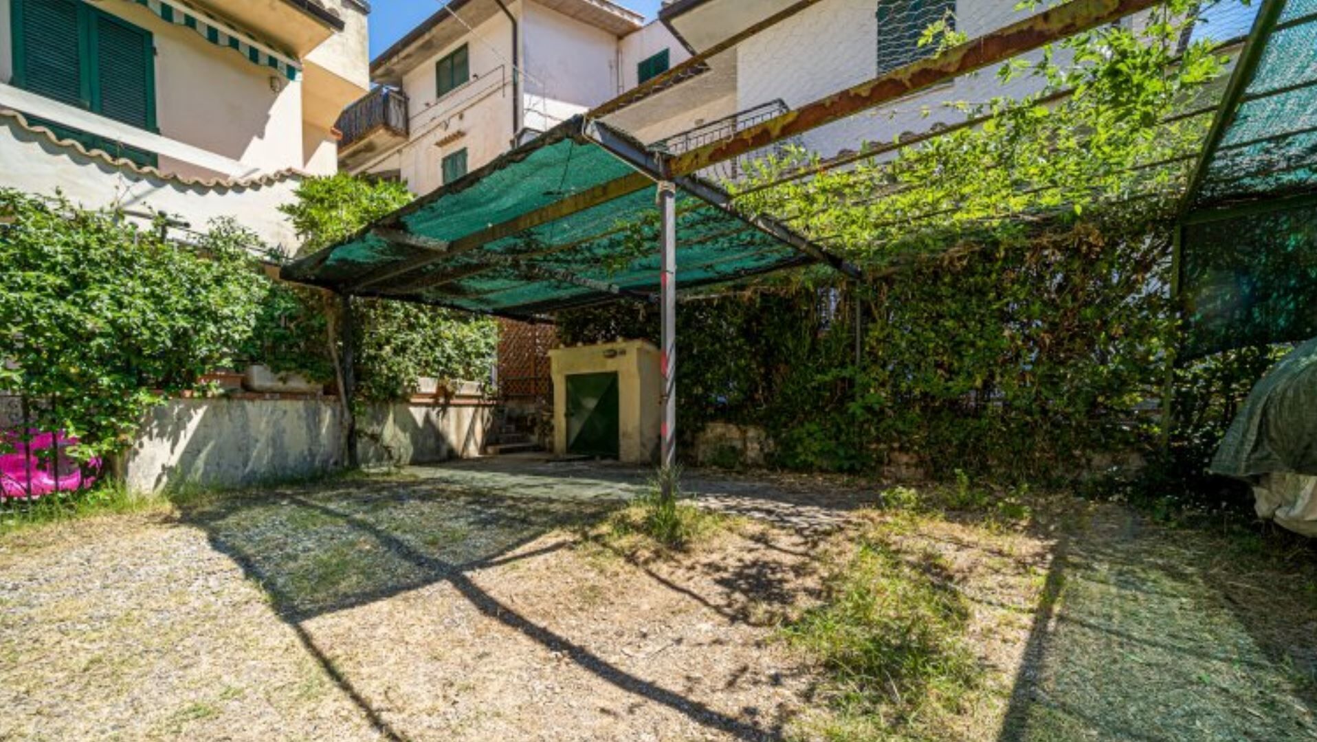 Un cortile con una pergola verde, un portoncino e un muro in cemento.