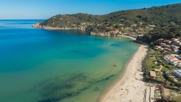 An aerial view of a coastal area with a clear beach, a small town with houses, and a hilly backdrop.