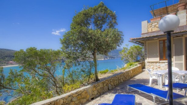 Un espace extérieur entouré de murs en pierre, offrant une vue sur la mer, un arbre et un bâtiment doté d’un balcon.