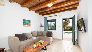 Un salon avec un plafond en bois, un canapé beige, une table basse en bois sur laquelle se trouvent une bouteille et des verres, et un téléviseur fixé au mur.