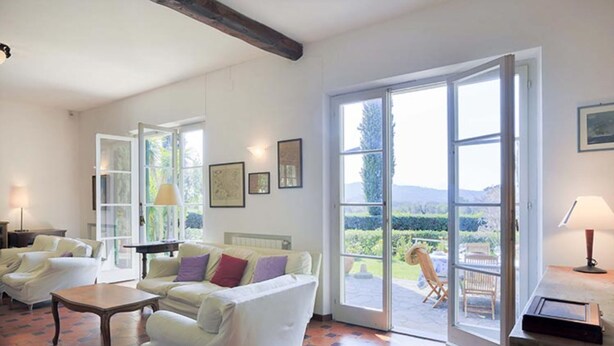 Ein geräumiges Wohnzimmer mit einem großen Schiebeglasfenster, das in einen Garten führt. Zwei weiße Sofas, ein Couchtisch und eine Lampe auf einem Beistelltisch sind vorhanden.