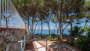 Un edificio de piedra con barandilla metálica que domina una zona costera con árboles y vista al mar.