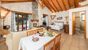 Une pièce chaleureuse avec une cheminée en pierre, des poutres apparentes en bois au plafond, un coin repas avec une table dressée pour deux personnes et un lit avec une table de chevet.