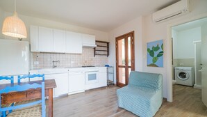 A compact kitchen with white cabinets, a built-in oven, and a dining area with a blue table and chair. - Casa al Mare in a panoramic position on the Gulf of Pareti (Capoliveri)