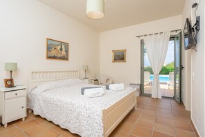 A bedroom with a bed, two bedside tables, a chair, a painting, and a view of an outdoor swimming pool.