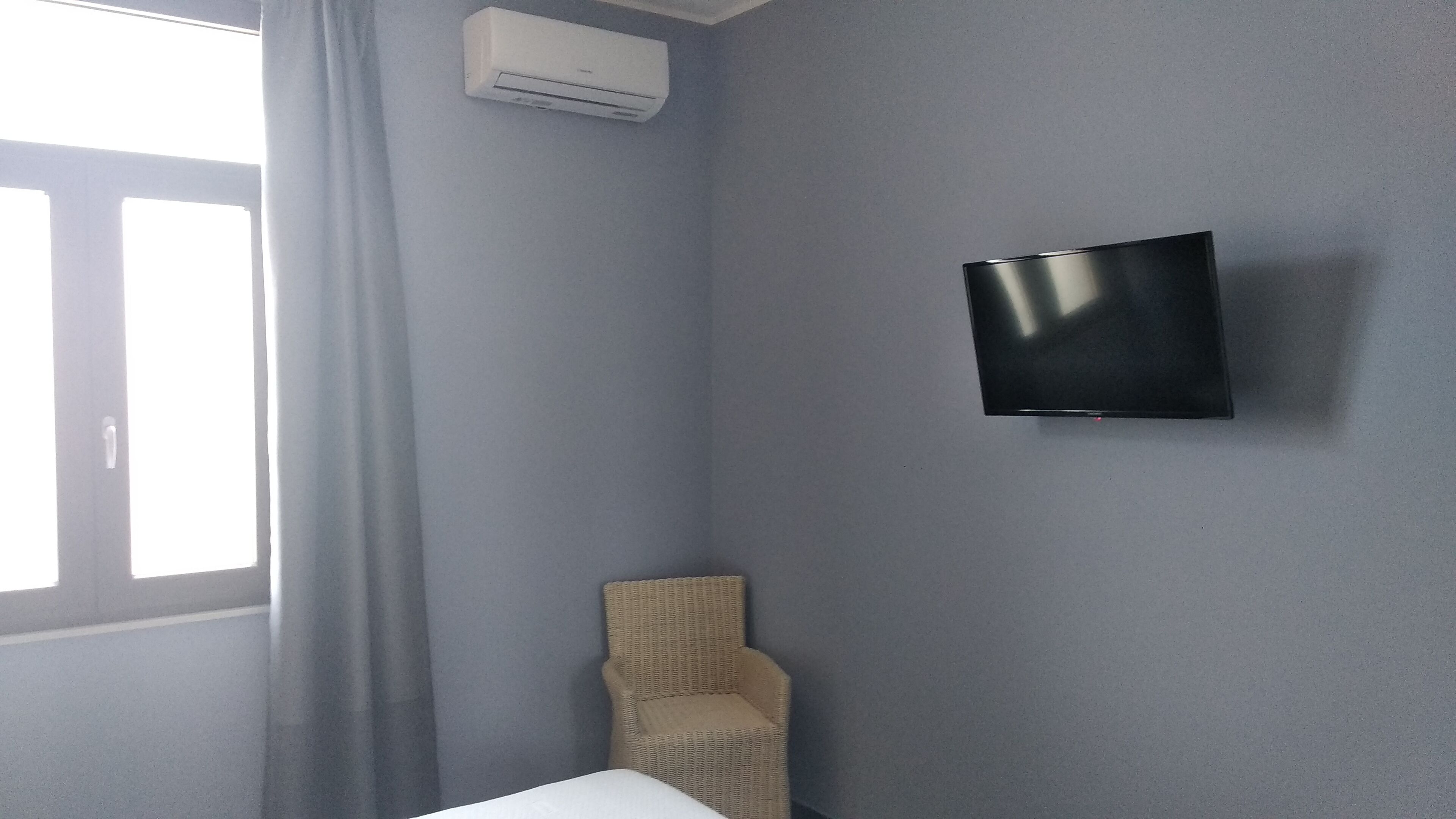 Una habitación con una cama individual, un televisor montado, una silla de mimbre y una ventana con cortinas.