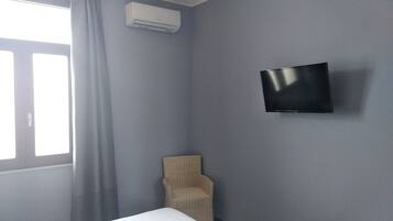 Una habitación con una cama individual, un televisor montado, una silla de mimbre y una ventana con cortinas.