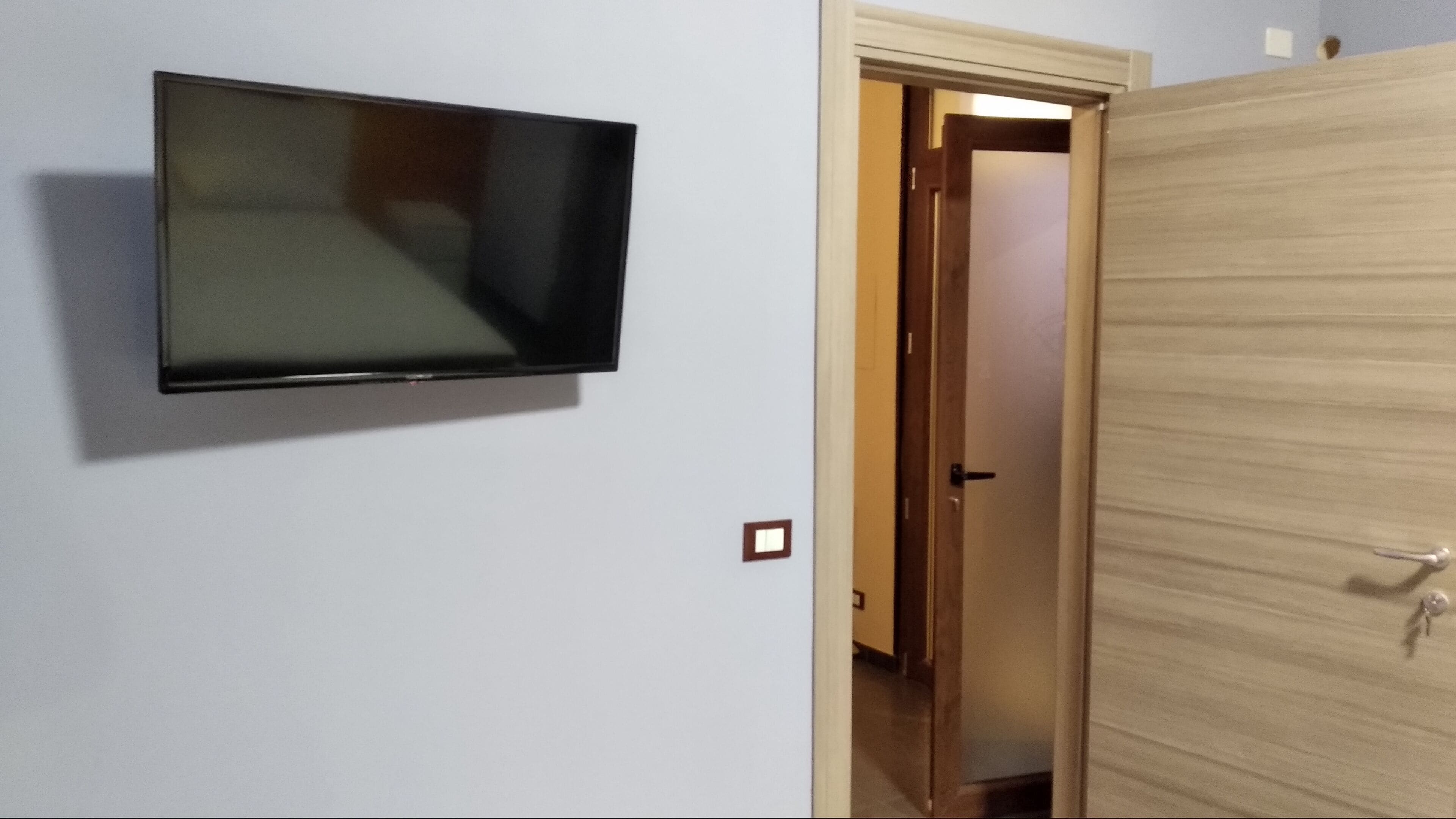 A room with a mounted television on the wall, a wooden wardrobe, and a door leading to another room.