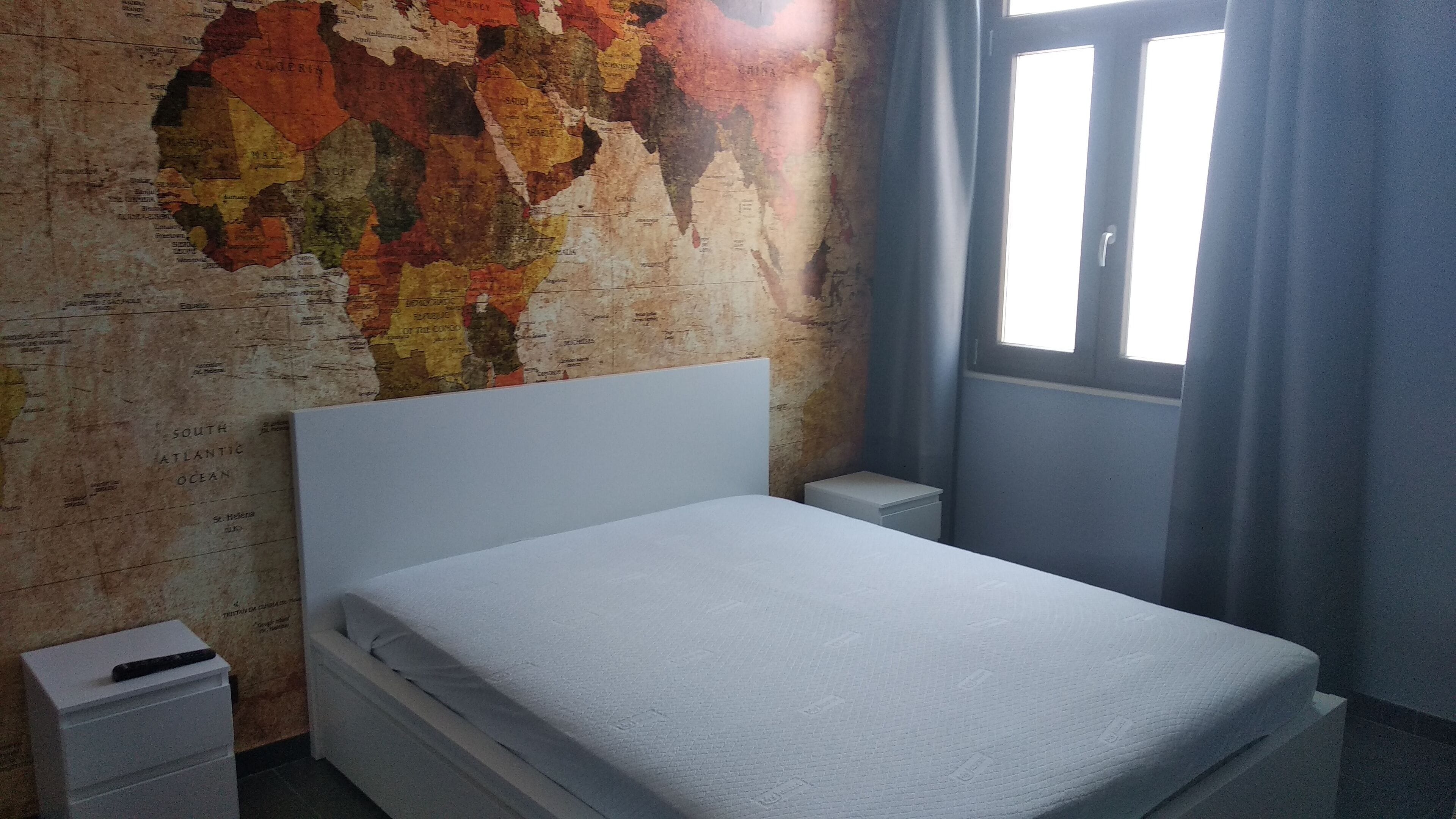 A room with a bed, a nightstand, a map on the wall, and a window with curtains.