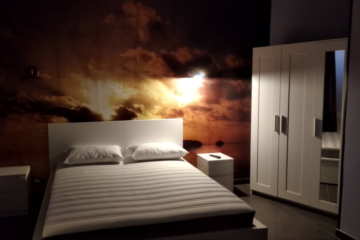 Ein Schlafzimmer mit Bett, Nachttischen, einem Kleiderschrank und einer Wandmalerei von einem Sonnenuntergang.