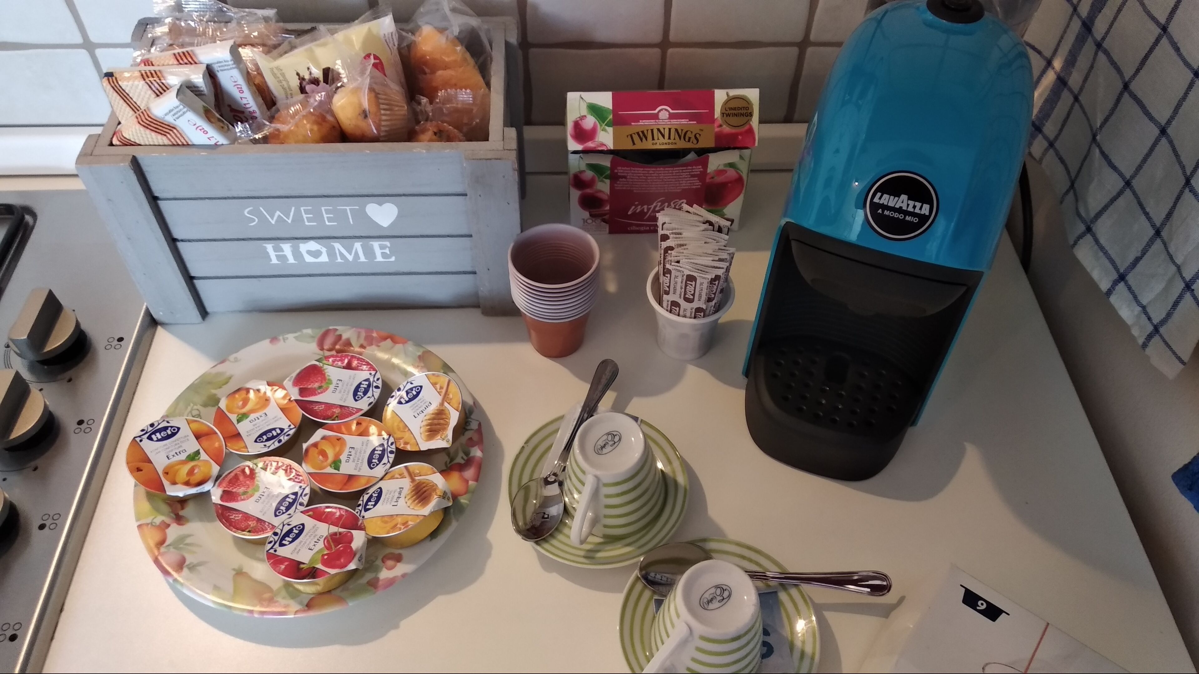 Un comptoir de cuisine avec un plateau de fruits, une boîte étiquetée « SWEET HOME », un distributeur d’eau et une machine à café.