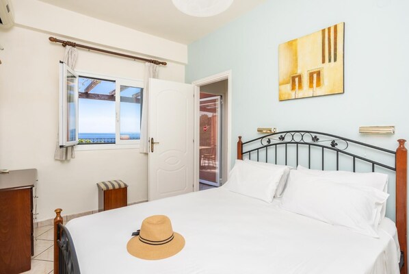 A bedroom with a bed, a window with a view, a door, a wall-mounted artwork, and a hat on the bed.