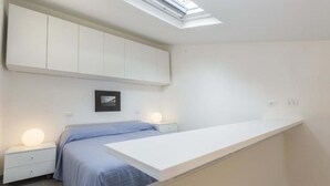 Ein Schlafzimmer mit Oberlicht, einem Bett mit blauer Bettdecke, zwei Nachttischen mit Lampen und einem weißen Kleiderschrank.