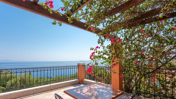 A terrace with a table and chairs, a pergola with pink flowers, and a view of the sea.