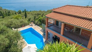 A two-story building with a swimming pool, surrounded by greenery and overlooking a large body of water.