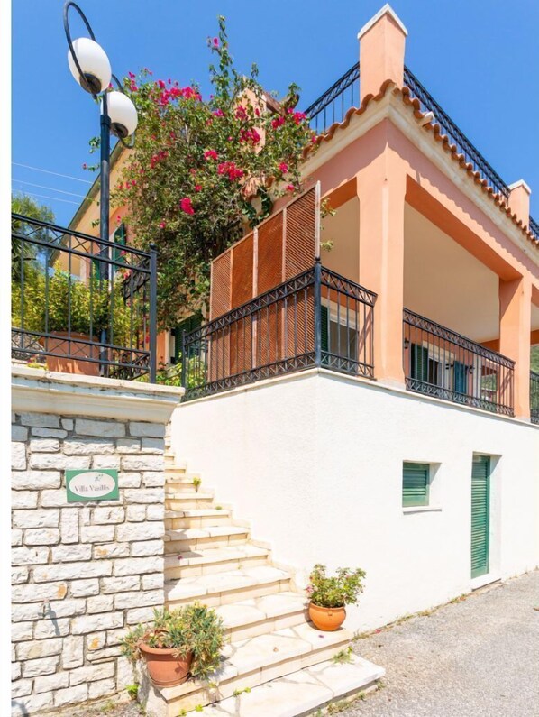 Edificio de dos pisos con entrada de muro de piedra, un balcón con barandas de metal y un letrero que indica 'Villa Vasillis'.
