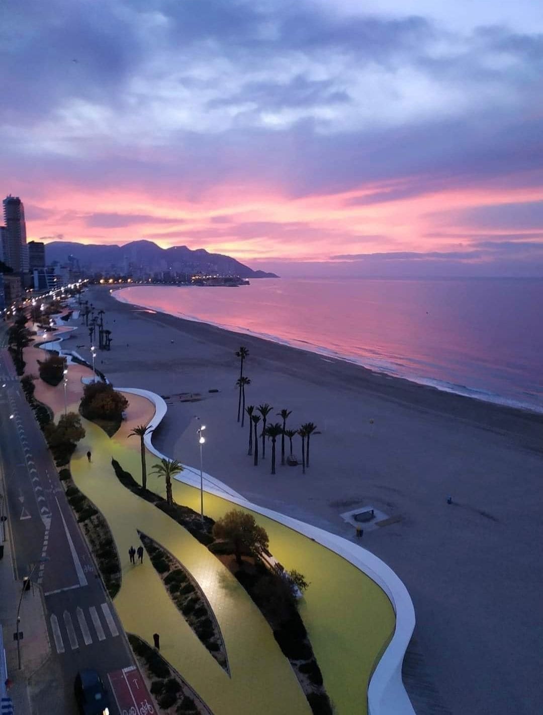 Un paisaje urbano costero al atardecer, con una rambla curva que conduce a una playa.