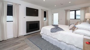 A modern hotel room with a large bed, a flat-screen TV mounted on the wall, a fireplace, and a view of the sea through the windows. - Newport Beach Modern Home steps to the Beach (Newport Beach)