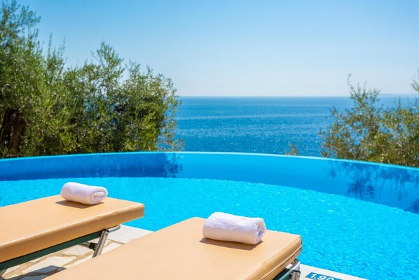 Una piscina infinita con dos tumbonas y toallas, rodeada de vegetación y con vistas al mar.
