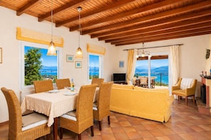 A room with a dining table, wicker chairs, a sofa, a TV, and a balcony with a view.