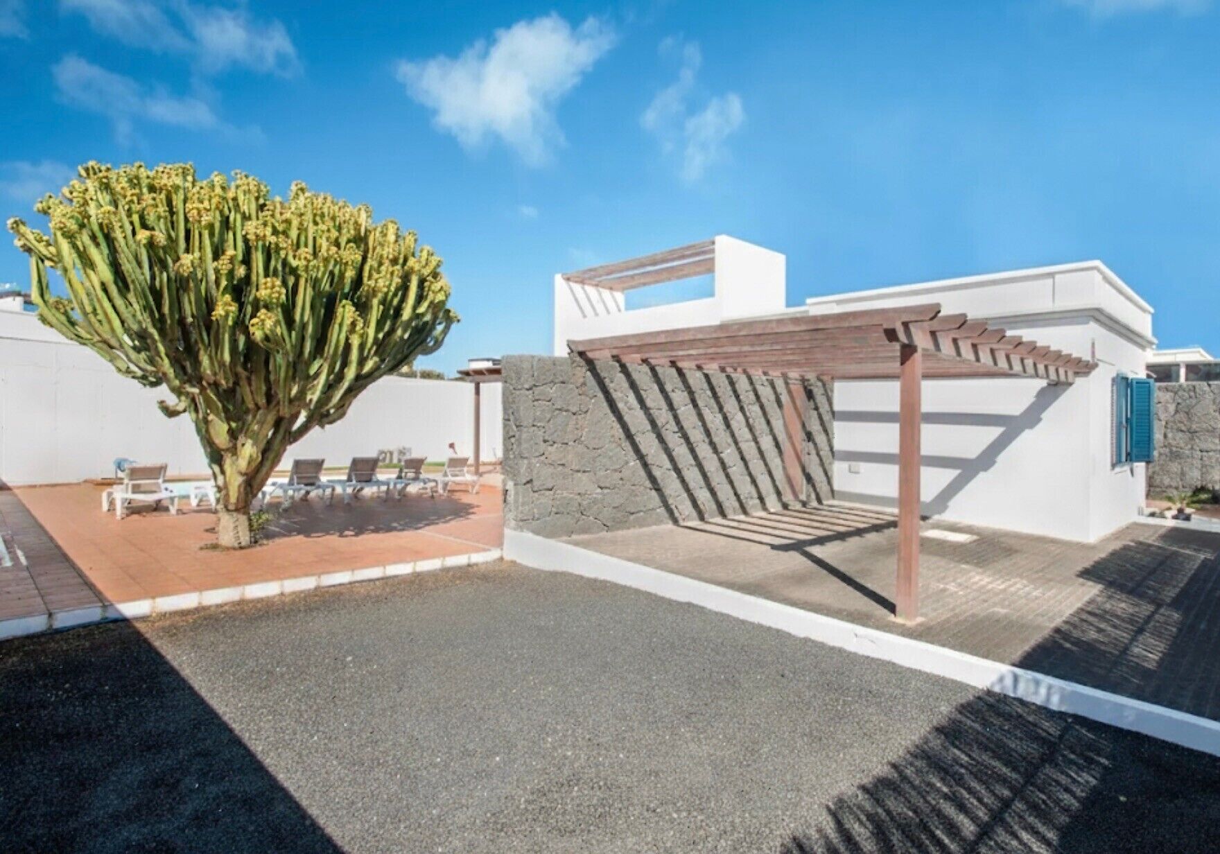 A modern outdoor patio with a pergola, a large cactus, and a white building with a stone wall.